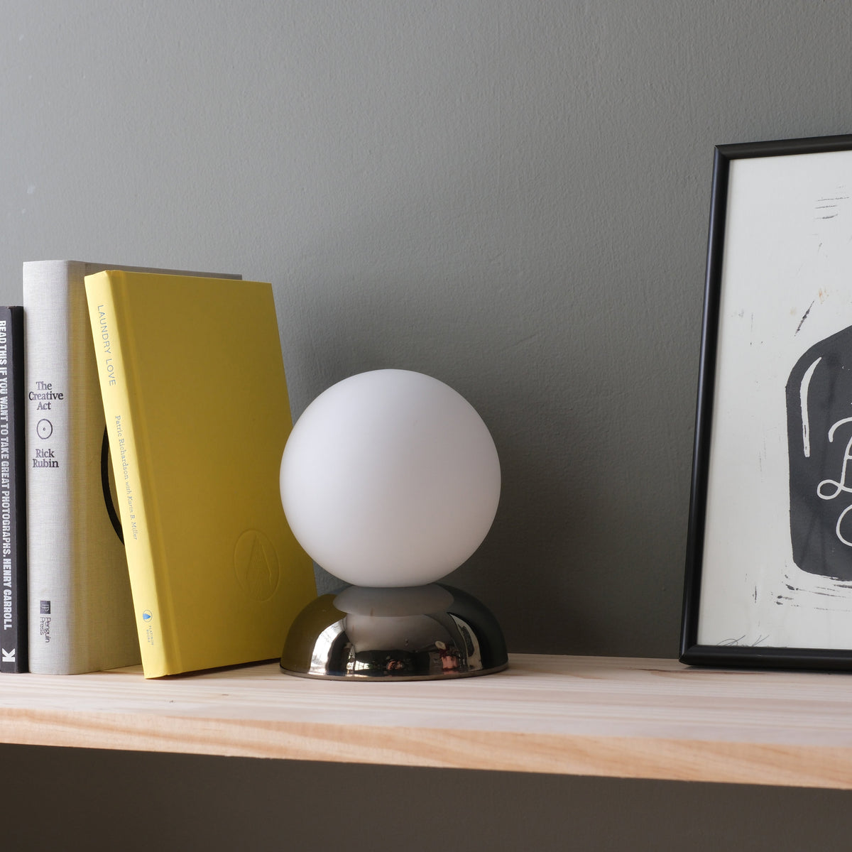 Small table lamp with a white spherical shade and reflective metallic dome base, placed on a light wood shelf alongside vertically stacked books and framed artwork.