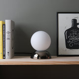 Spherical white lamp with a reflective metallic dome base on a light wood shelf, flanked by vertically aligned books on the left and a framed black-and-white print on the right.