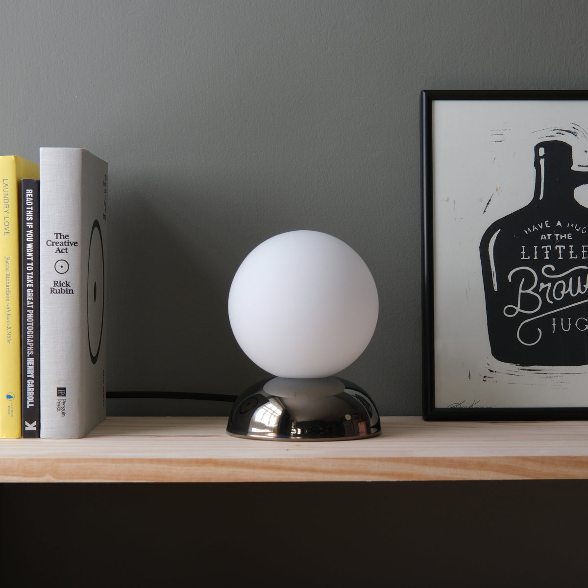 Spherical white lamp with a reflective metallic dome base on a light wood shelf, flanked by vertically aligned books on the left and a framed black-and-white print on the right.