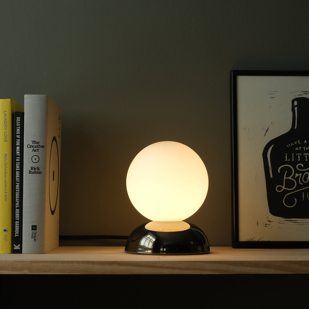Glowing spherical white lamp on a glossy black rounded base, placed on a wooden surface between colorful books and framed artwork against a dark wall.