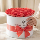 Round white box filled with tightly arranged coral roses and wrapped with a matching coral ribbon tied in a bow, held by a hand with nude nail polish.