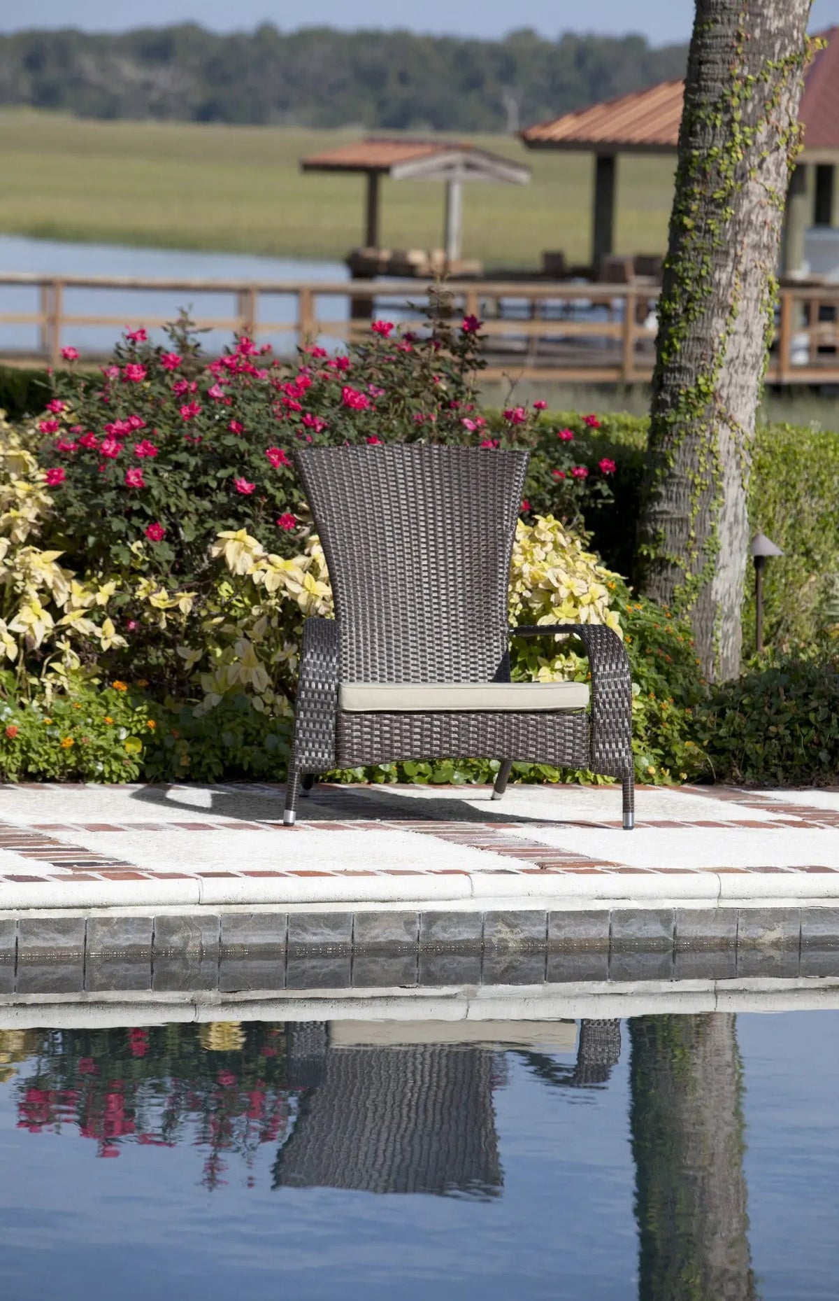 Brown synthetic wicker chair with beige cushion sitting on poolside patio tiles, reflected in water, with flowering bushes, tree trunk, and lake in background.