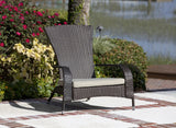 Dark brown woven wicker outdoor chair with a beige seat cushion, placed on a patio with red brick accents near colorful bushes and a waterfront background.
