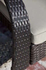 Close-up of dark brown woven wicker armrest and base with light beige cushion on a Coconino chair, casting soft shadow on a patterned outdoor surface.