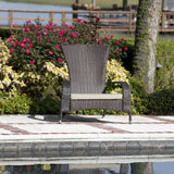 Single dark brown woven wicker armchair with beige cushion on a tiled patio beside a pool, backed by green and flowering shrubbery and a wooden fence.