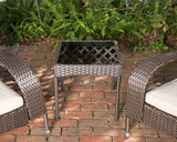 Square wicker patio side table with black glass top between two curved armchairs with white cushions on a brick patio with green plants in the background.