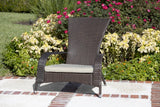 Dark brown wicker outdoor chair with curved arms and a light gray cushion, positioned on a stone patio bordered by red and white brick accents and colorful flowering shrubs.