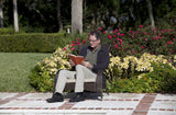 Man sitting on dark brown wicker chair reading an orange book outdoors, with green grass, yellow and red flowers, and trimmed hedges in the background.