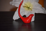 Small red bird-shaped ring holder with a long curved tail on a dark surface, positioned in front of a large white flower with a yellow center.