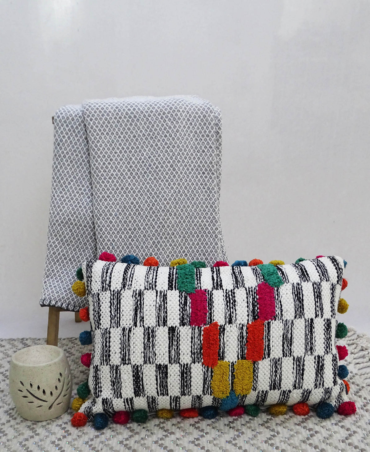 Rectangular handwoven throw pillow with black and white checkered pattern and colorful pom-poms along edges, placed on patterned light gray fabric draped over wooden chair legs.