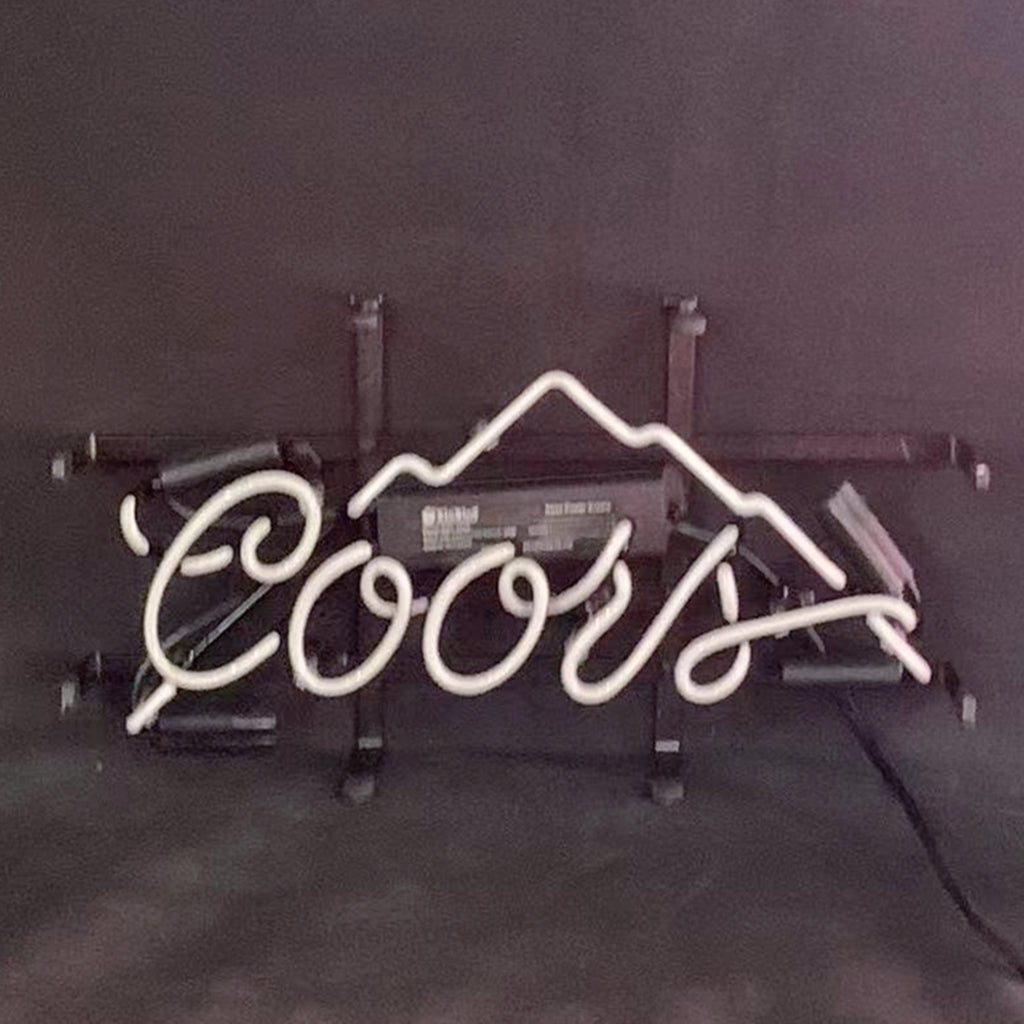 White neon sign spelling 'Coors' with a mountain outline above the text, mounted on a black frame against a dark background.