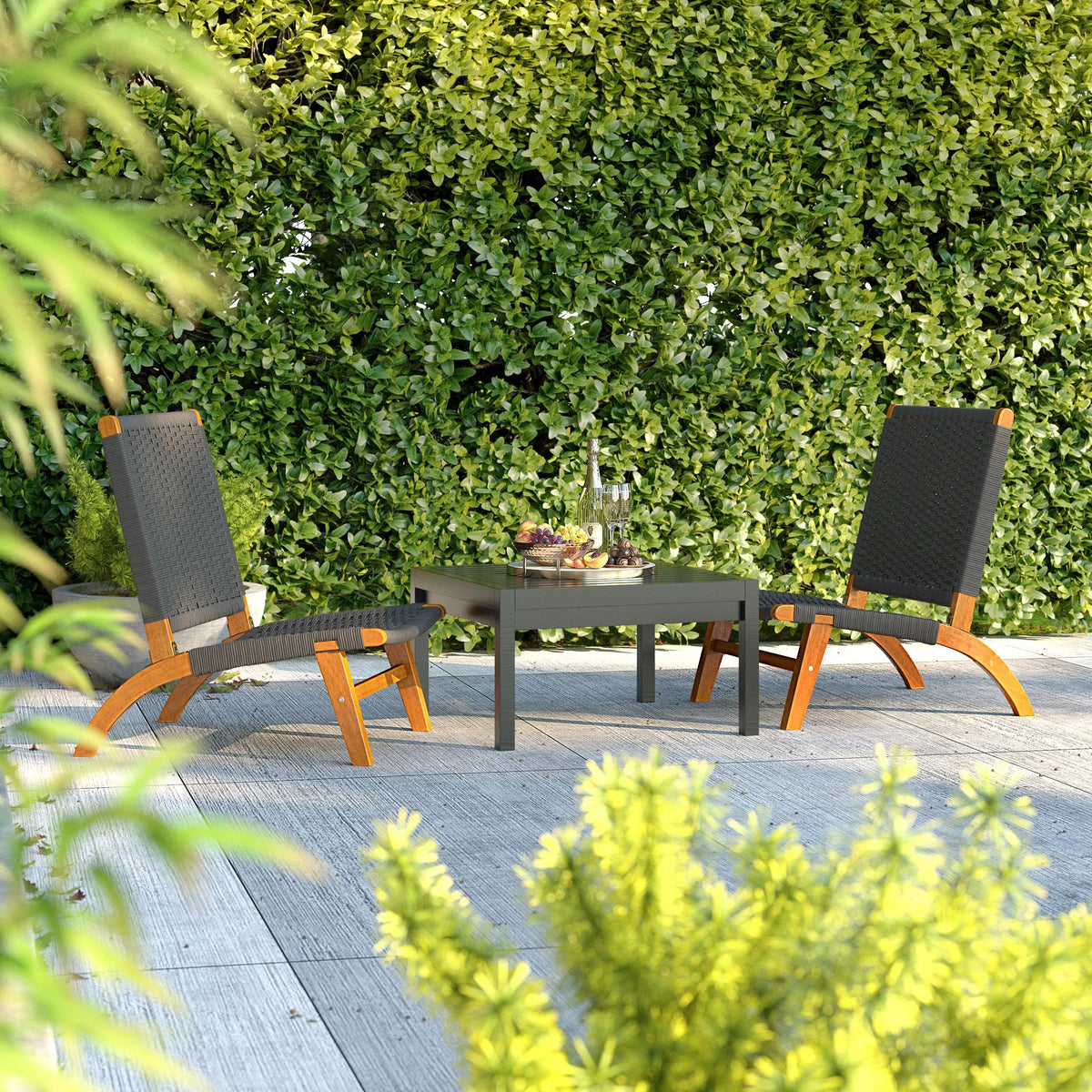 Two modern lounge chairs with curved wooden frames and black woven seats placed around a square black table on a gray tiled patio, with dense green foliage background.
