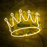Yellow neon sign shaped like a crown mounted on a clear rectangular panel, hanging against a textured brown wall with visible electrical components behind.