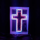 Neon sign shaped like a cross with a glowing pink interior outline and a blue outer outline, mounted inside a clear acrylic box against a dark background.