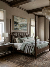 Bedroom with a dark wood four-poster bed made up with layered light grey and green bedding, flanked by matching nightstands and soft beige walls, beneath a large abstract painting.