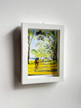 Framed watercolor painting of bikers cycling along a sunlit tree-lined path by the Hudson River, with green and yellow foliage and blue sky patches.