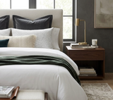 Bed with black and white patterned pillows, textured beige lumbar pillow, and dark green throw, next to a wooden nightstand holding books and a brass lamp