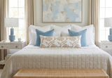 Bed with white linens, beige quilt, two blue square pillows, and a long beige floral-patterned lumbar pillow, flanked by light wood nightstands with blue lamps.