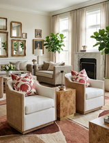 Living room with two white cushioned armchairs featuring Sunbaked Red desert floral pillows, a burl wood side table between them, beige sofa and large green plants by tall windows.