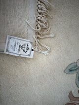 Close-up of a cream-colored hand-tufted wool rug corner with knotted fringe and a white label attached, showing part of a floral design in muted green and brown tones.