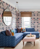 Living room with blue textured L-shaped sofa and multicolored pillows against wallpaper featuring abstract circular patterns in navy, pink, and beige.