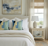 Bedroom with white bedding and two square pillows featuring abstract blue, green, yellow, and black geometric patterns, complemented by beige curtains and a carved wood nightstand.