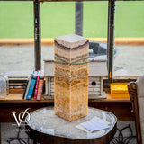 Tall square lamp with layered brown, beige, and green stone resembling onyx, placed on a round glass-top table beside books and a white birdhouse near a window.