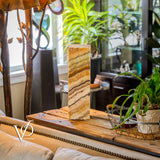 Tall square table lamp with layered amber and cream onyx stone base sitting on a rustic wood console table surrounded by green plants and a metal tree lamp.