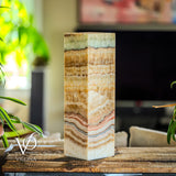 Tall square table lamp with layered bands of aqua, brown, beige, and coral hues, standing on a rustic wooden surface with plants nearby and a blurred background.