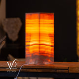 Rectangular onyx table lamp glowing with warm orange and red horizontal bands of varying opacity, placed on a wooden surface against a dark background.
