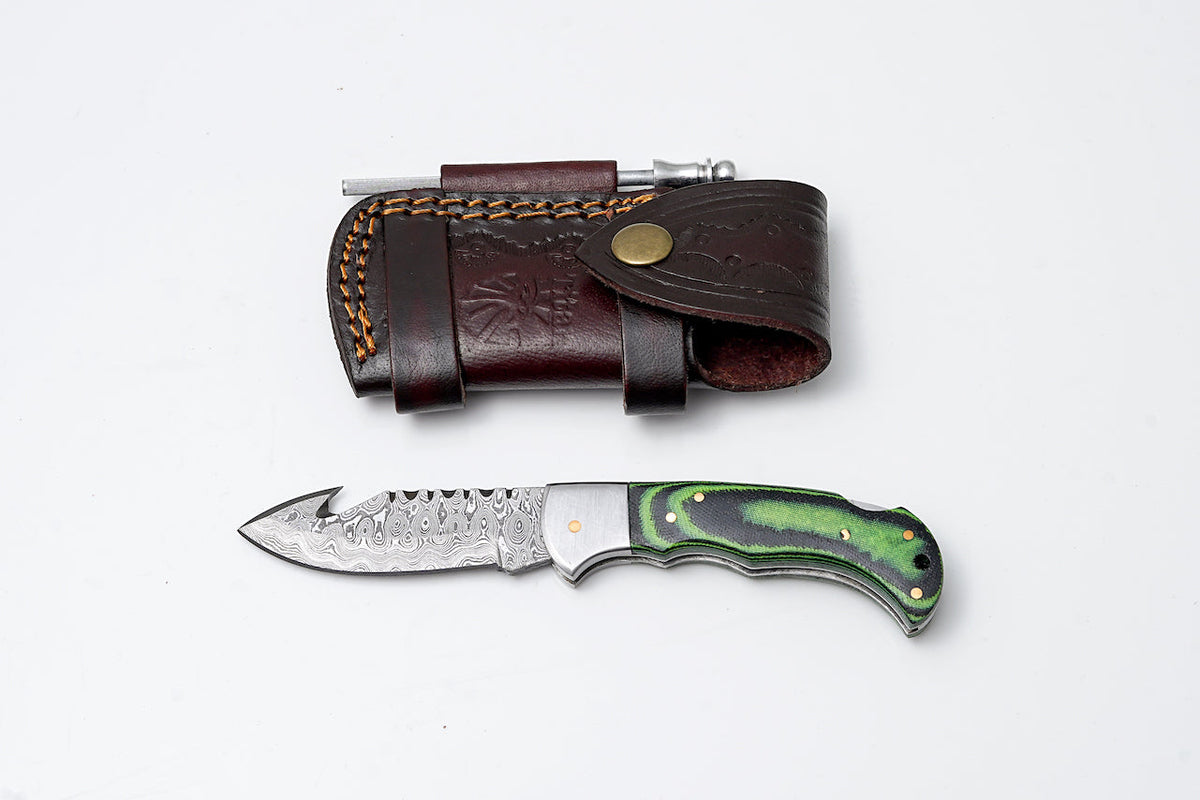 Green Micarta Damascus Gut Hook Hunting Knife TF-103 with leather sheath and fire starter laid on wood surface