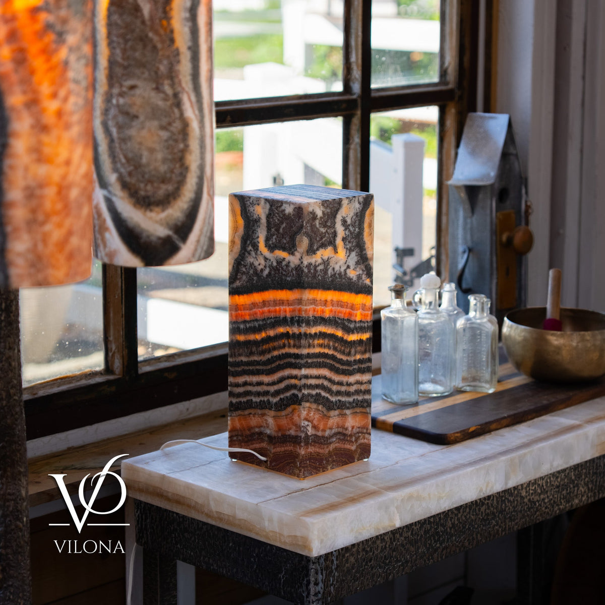 Rectangular onyx table lamp with warm orange and brown layered patterns glowing softly on a white marble surface near a window with glass bottles and wooden objects.