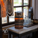 Rectangular block lamp with distinctive black, brown, and vibrant orange tangerine onyx patterns, placed on a white marble-top table by a window with clear glass bottles nearby.
