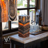 Rectangular table lamp with orange and black striped pattern, resembling onyx stone, placed on a marble-topped table by a window with vintage glass bottles and rustic decor.