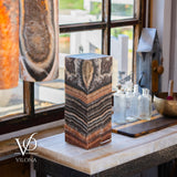 Rectangular block of tangerine-colored onyx with layered orange, brown, and cream patterns standing upright on a marble countertop near a rustic window with vintage glass bottles.
