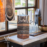 Rectangular onyx table lamp with layered black, brown, and beige stone patterns on a white marble countertop by a window with glass containers and brass bowl nearby.