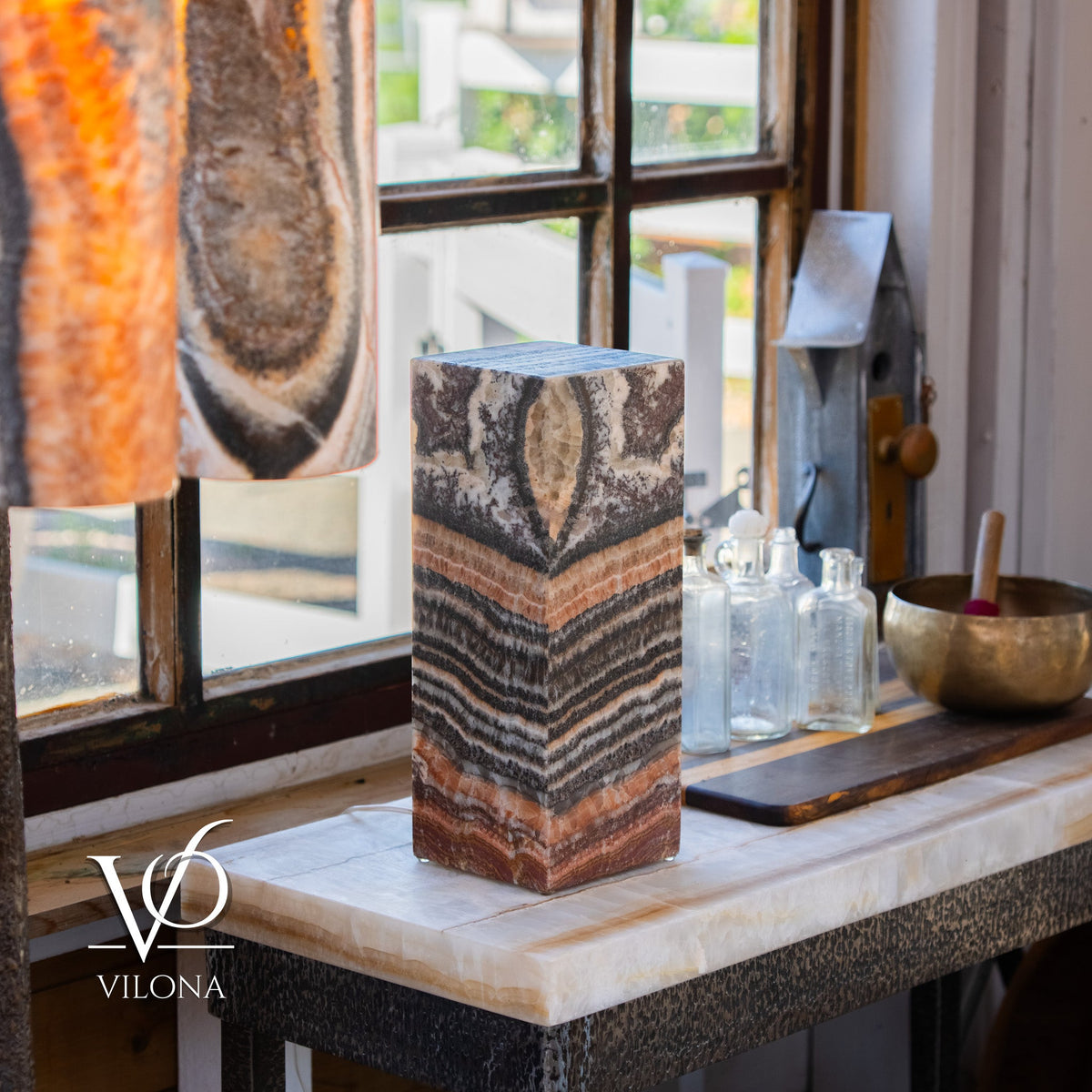 Rectangular tall lamp made of tangerine and black onyx with natural striped and circular patterns, placed on a windowsill alongside glass bottles and a brass bowl.