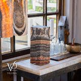 Tall rectangular onyx table lamp with natural horizontal bands of brown, black, and cream, displayed on a marble countertop by a window with glass bottles and a brass bowl nearby.