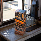 Tall rectangular table lamp with vibrant orange and black onyx patterns glowing through translucent stone, placed on a light marble surface by a window with vintage glass bottles