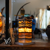Rectangular tangerine onyx table lamp with glowing horizontal orange, white, and black layers, placed on a book on a rustic wooden surface indoors.