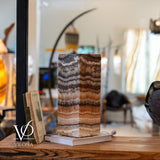Rectangular tangerine onyx table lamp with horizontal banding in shades of orange, brown, and cream, placed on a wooden surface with books and blurred background.