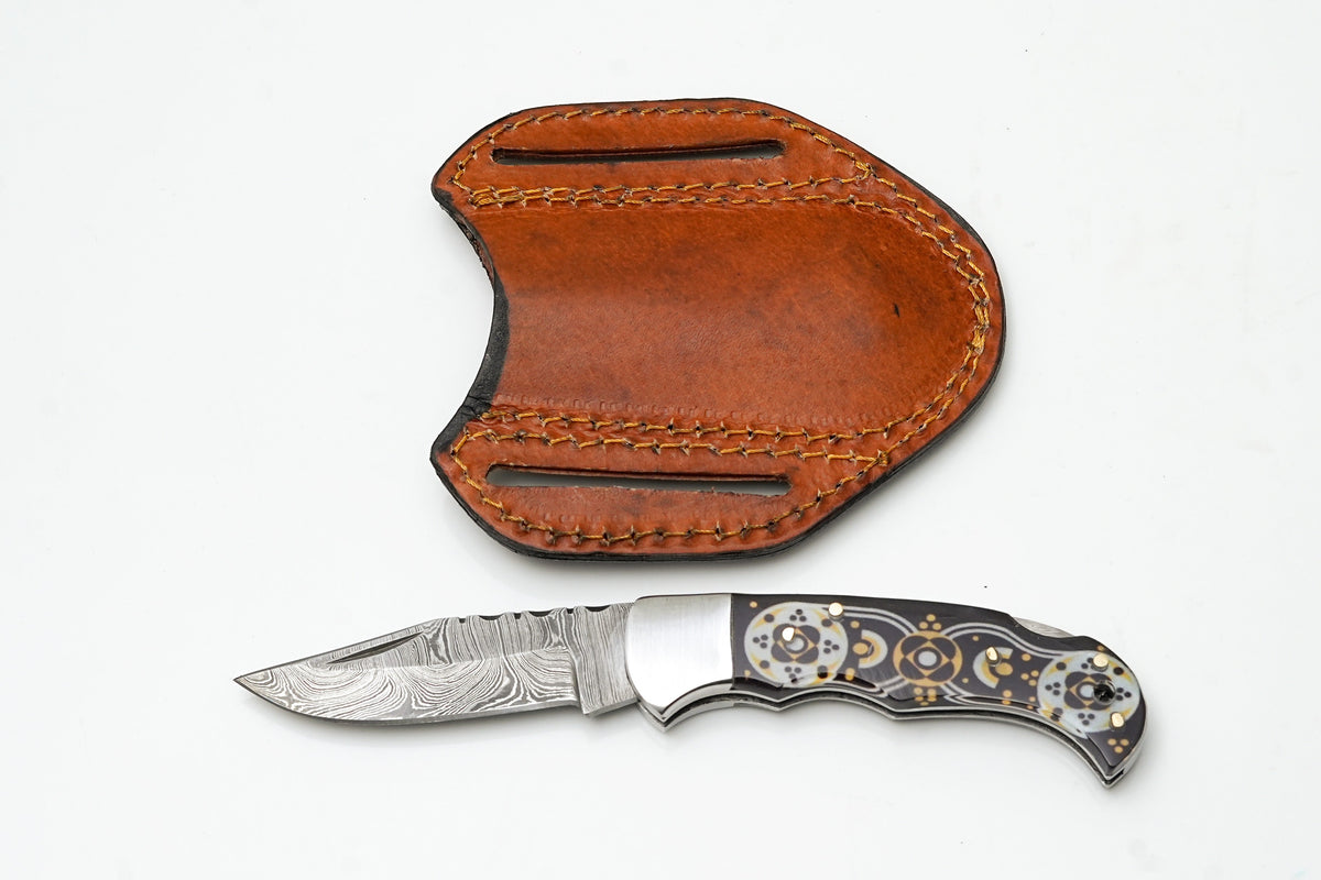 Hand-Forged Damascus Drop Point Folding Knife with black and gold mosaic handle resting on a cloth surface alongside a leather pancake sheath.