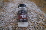 Clear plastic bottle labeled 'Cherry Rush Corn Dust' with white powder inside, lying on a bed of kernels with a beige and brown color palette.