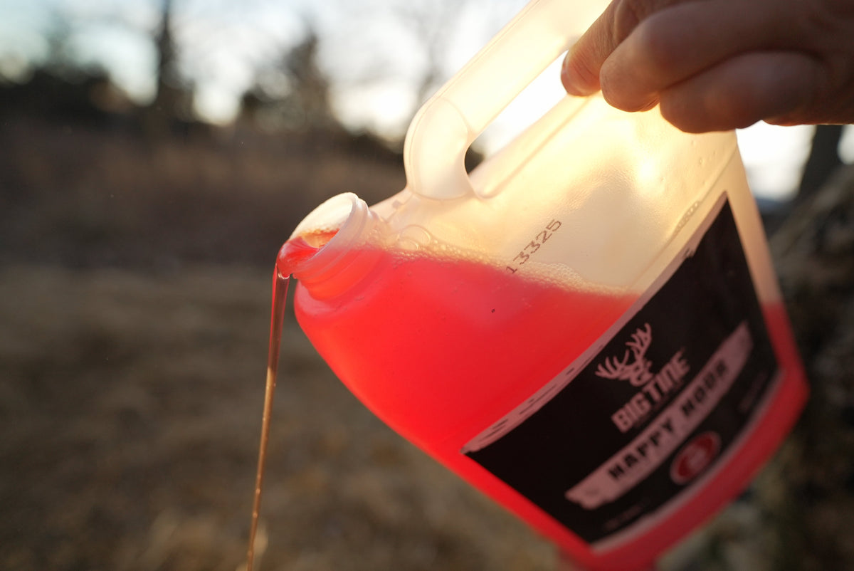 Close-up of a translucent red liquid being poured from a white plastic container labeled with a deer antler logo and the words 'Big Tine Happy Hour' outdoors.