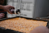 Close-up of a hand sprinkling a container of Corn Dust seasoning over a large pan filled with yellow corn kernels, with a blurred background showing another person and containers.