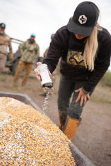 Person wearing black cap and hoodie shaking white salt shaker over a large pile of yellow and white cracked corn outdoors with three blurred people in the background.