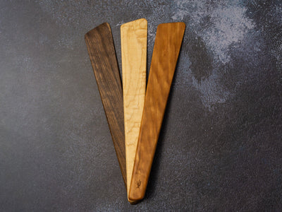 Wooden spatula resting on a dark textured surface, showing wood grain and slender shape
