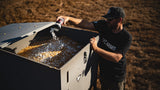 Person wearing a black T-shirt and cap pouring white powder from a container into a large open gray metal box filled with corn kernels outdoors on dry grass.