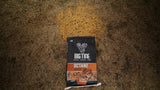 Black and orange bag of Big Tine Octane attractant with scent label, placed on dry grass with a noticeable circular patch underneath.