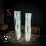 Pair of tall, hexagonal white onyx floor lamps with vertical brown veining standing on a textured rug in a dim room with dark walls.